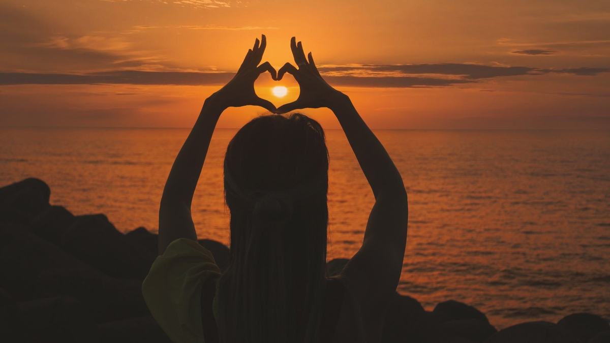 A person silhouetted against a sunset over the sea, hands raised to frame the sun in a heart shape.