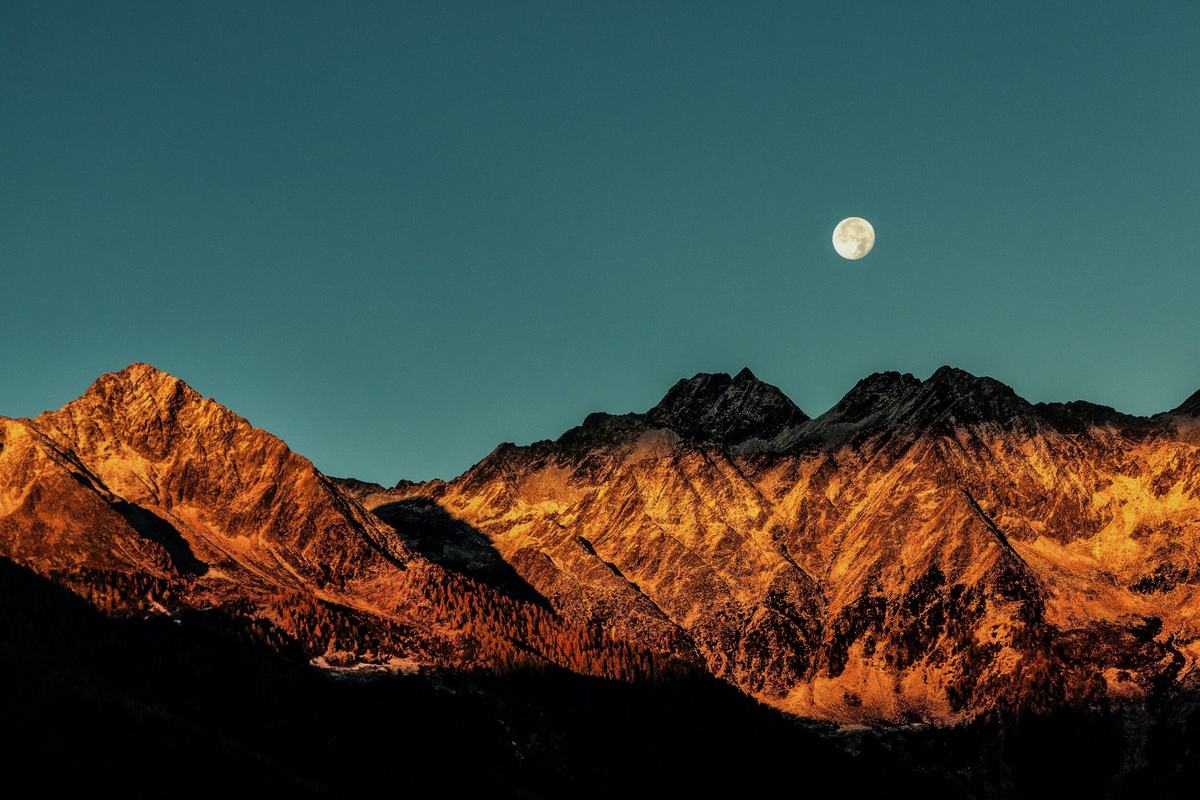 A full moon in a deep blue sky above mountain peaks lit orange by the setting sun.