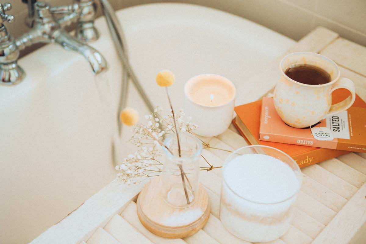 A bath tray holding a lit candle, a mug of tea, books and a glass of milk.