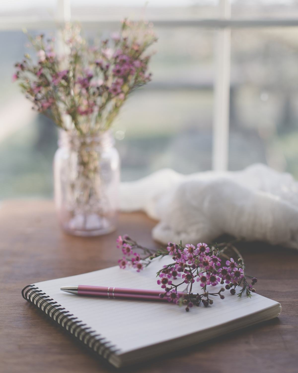 An open spiral notebook and a pink pen on a wooden table by a window, with wax flowers in a jar.