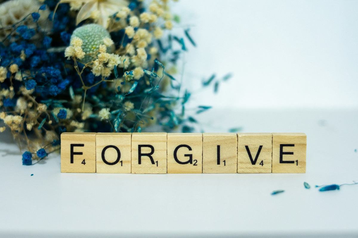 Scrabble tiles on a white surface spelling the word FORGIVE, beside dried blue flowers.